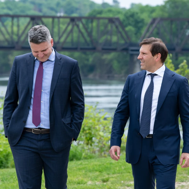 Gideon Asen attorneys Ben Gideon and Taylor Asen walking together along a Maine riverbank with a historic iron bridge in the background