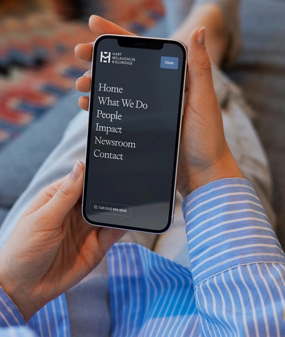 erson holding a smartphone displaying the Hart McLaughlin & Eldridge mobile navigation menu with links to Home, What We Do, People, Impact, Newsroom, and Contact