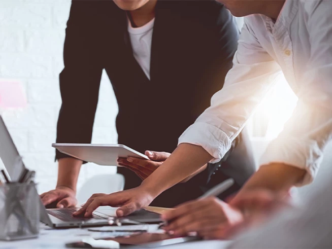 Close-up of coworkers collaborating at a desk—one typing on a laptop, another holding a tablet, with papers and pens on the table and warm sunlight streaming in from the right.